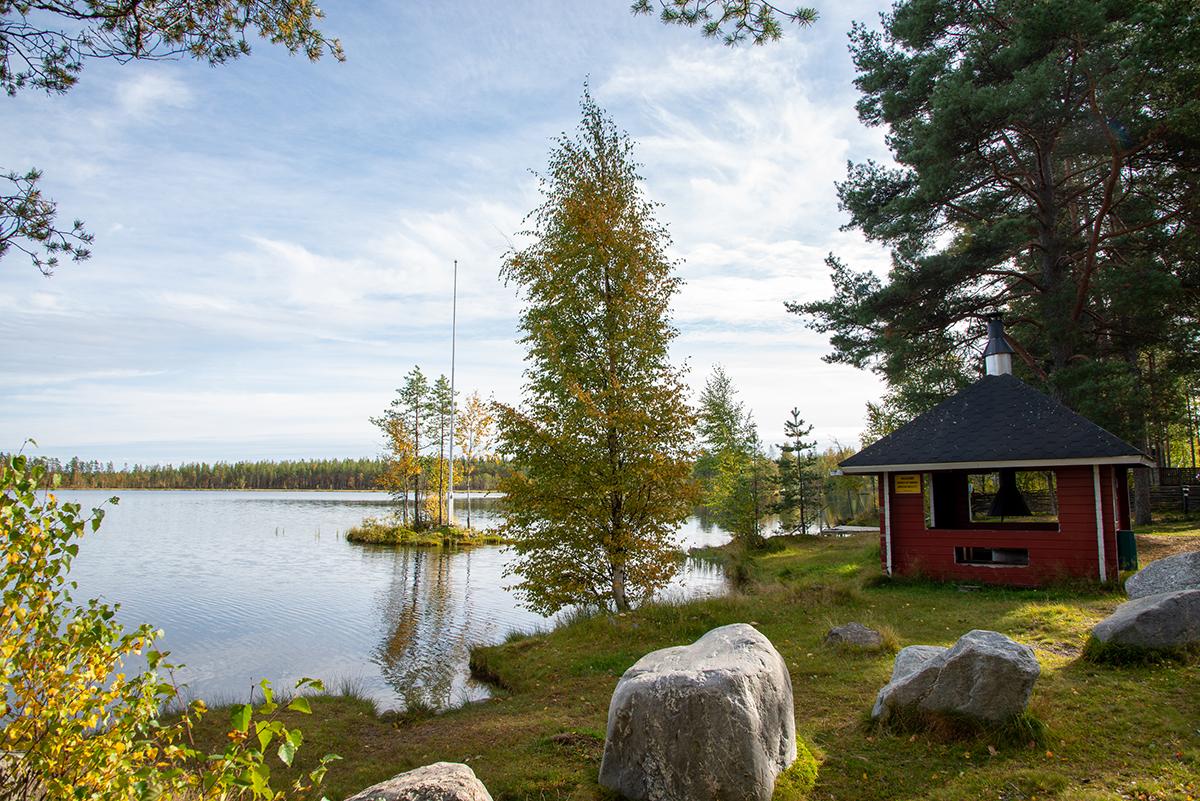 Hiukkajärven ranta ja grillikatos. Kuvassa etualalla isoja kiviä, punainen hirsinen grillirakennus, koivuja ja järvessä pieni saar, jossa lippumasto.