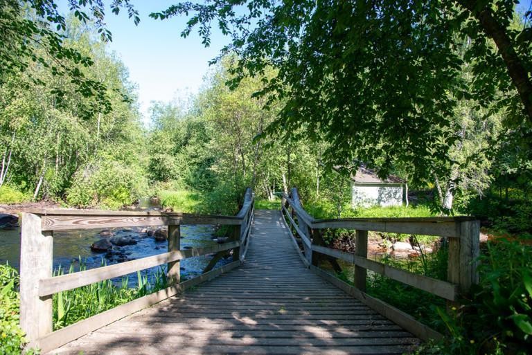 Kesäisessä maisemassa puinen silta vie joen yli. Kuva melko varjoisa.