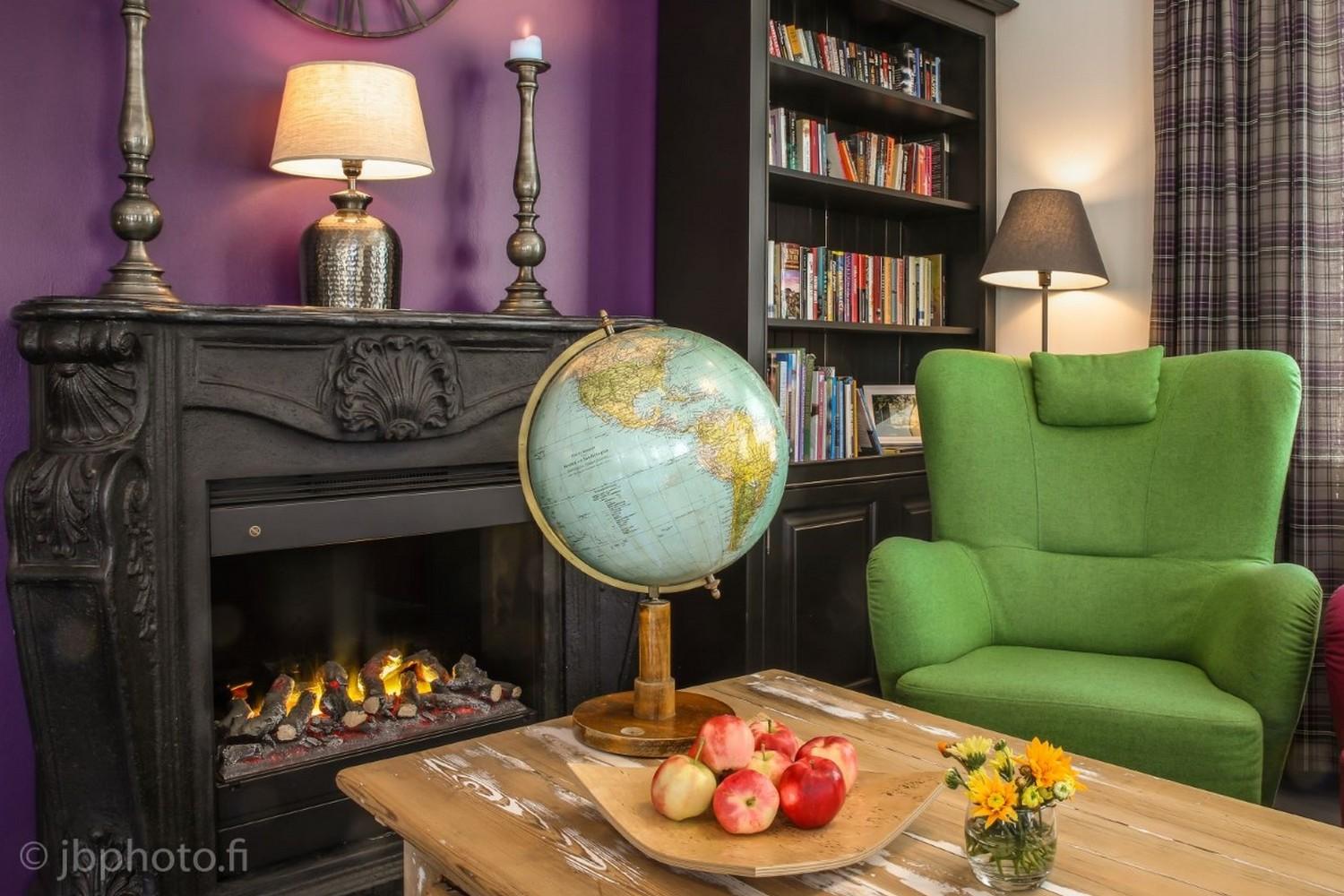 Kuva Hiljahelenan oleskelutilasta. Etualalla pöytä, jolla karttapallo, omenavati ja kukkamaljakko, taustalla kirjahylly, takka, lamppuja ja nojatuoli.