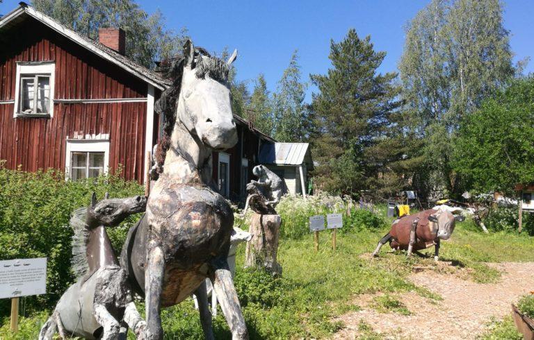 Etualalla kaksi pääosin metallista valmistettua hevoshahmoa, kauempana ruosteisen näköinen lehmähahmo, taustalla punainen rakennus.