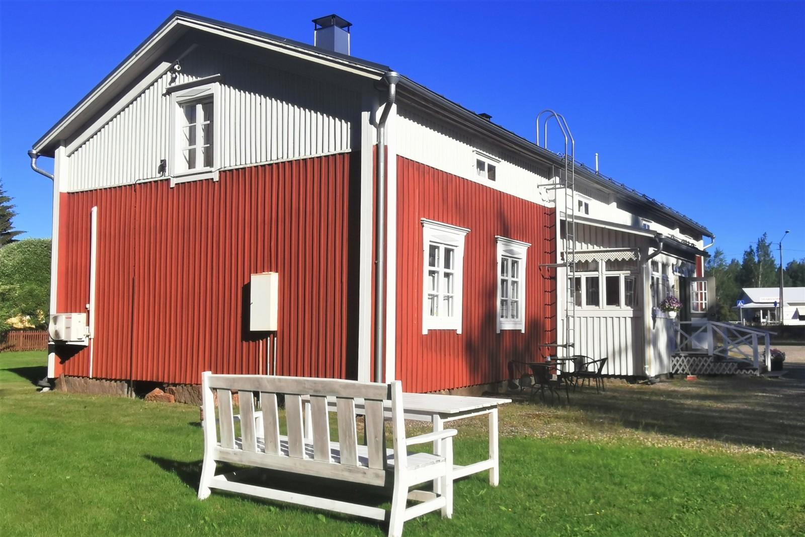 Punaninen puinen puolitoistakerroksinen talo, etualalla valkoinen penkki ja pöytä.