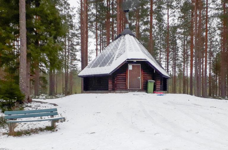 Talvikuvassa keskellä kota, etualalla sininen penkki ja taustalla mäntyjä.