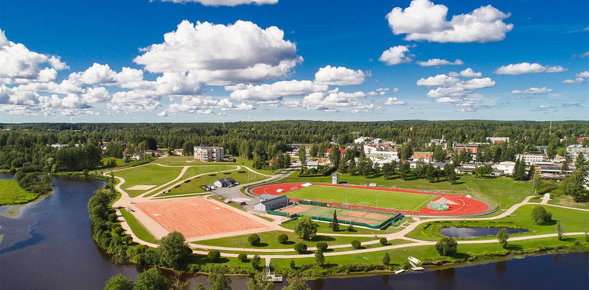 Kesäinen ilmakuva Kurikan kaupungista, etualalla joki jonka takana urheilupuisto. Taustalla rakennuksia ja puita, sininen pilvinen taivas.