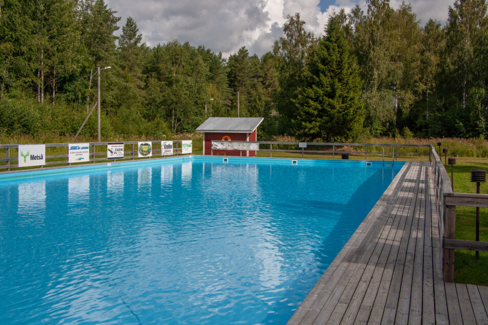 Maauimalan uimalassa. Puinen aita ja uima-allas on puisen terassin keskellä. Taustalla sekametsää ja punainen rakennus, jonka seinässä on pelastusrengas.