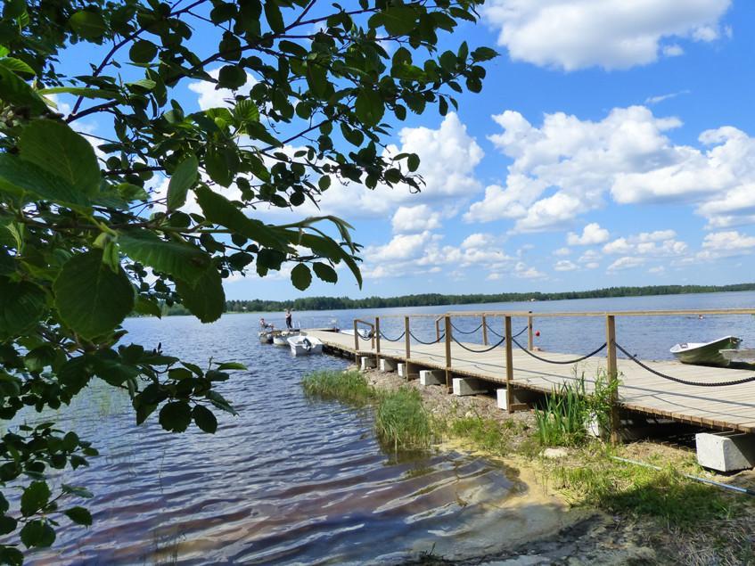 Pitkä puinen laituri järven rannassa, jossa on köysikaiteet. Taka-alalla sininen pilvinen taivas.