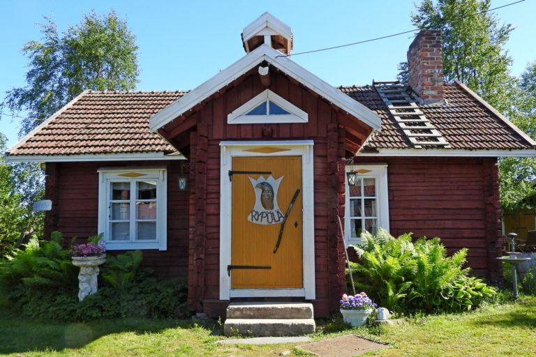 Kesäisessä kuvassa vanha punainen hirsirakennus, edestä päin kuvattuna. Keltainen ovi, jossa torpan nimikyltti.