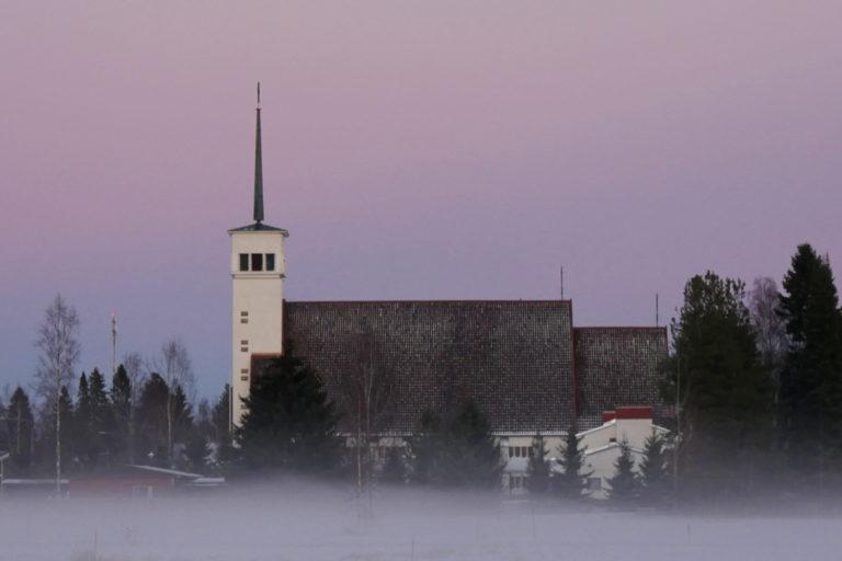 Teuvan kirkko kuvattu sivusta, usvainen talvikuva, taivas on värjäytynyt violetiksi.